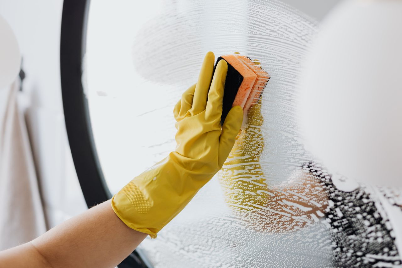 Une personne anonyme en gants en caoutchouc jaunes utilise une éponge simple pour nettoyer un miroir dans une salle de bain