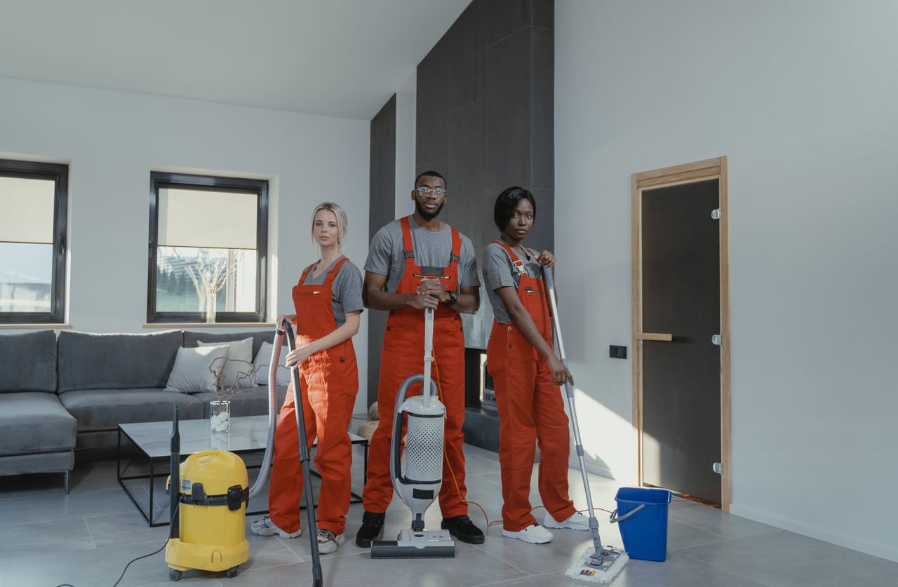 Équipe de nettoyage variée en uniformes rouges, prête à assainir un salon moderne avec aspirateur et serpillière.